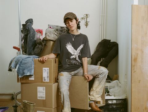 Portrait of Ju Aichinger sitting on cardboard boxes, surrounded by shoes and clothes.