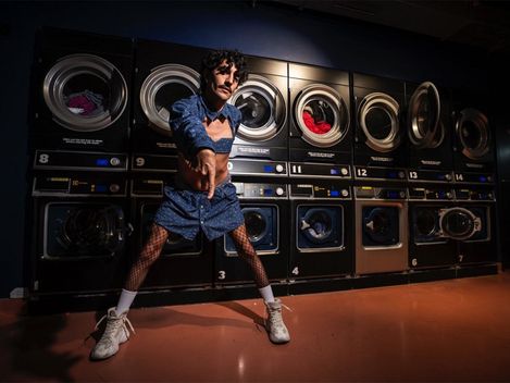 Man with mustache and blue shirt stands with legs wide apart in front of a row of washing machines in a laundromat pointing forward with his hand