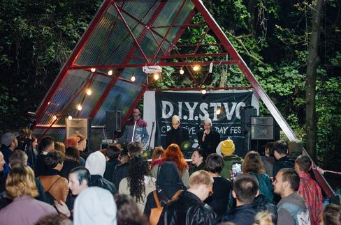 Crowd in front of a small stage with a triangular roof and a banner reading "D.I.Y.STVC" in a wooded outdoor area.