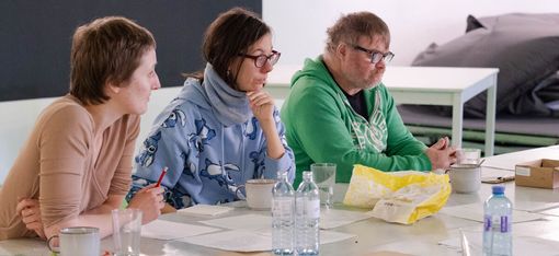 Drei Personen sitzen an einem Tisch mit Papieren, Flaschen und Tassen, eine Person trägt eine blaue Jacke mit Muster, eine andere eine grüne Kapuzenjacke.