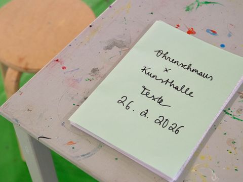 Notepad with handwritten text 'Ohrenkmaus x Kunsthalle Texte 26. 2. 2026' on a paint-splattered table with a wooden stool on a green floor in the background