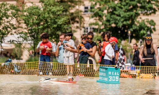 Mehrere Personen mit Kindern stehen hinter einem Absperrzaun an einem Wasserbecken im Haupthof des MuseumsQuartier, auf dem ein Modellboot mit einer Flagge und der Aufschrift 'SUPER AWESOME REAL 305' schwimmt
