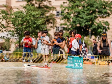 Mehrere Personen mit Kindern stehen hinter einem Absperrzaun an einem Wasserbecken im Haupthof des MuseumsQuartier, auf dem ein Modellboot mit einer Flagge und der Aufschrift 'SUPER AWESOME REAL 305' schwimmt