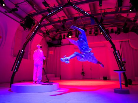 In einem Trainingslabor für Astronaut*innen: Die in weiß gekleidete Person, die von hinten zu sehen ist, spielt ein Instrument. Die Person in Ausbildung macht einen großen Sprung und schwebt in der Luft.