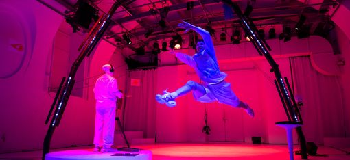 In a training laboratory for astronauts: the person dressed in white, seen from behind, is playing an instrument. The trainee takes a big leap and floats in the air.