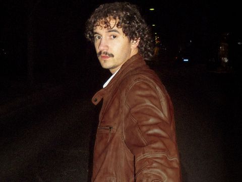 Person with curly hair wearing a brown leather jacket standing sideways on a dark street.