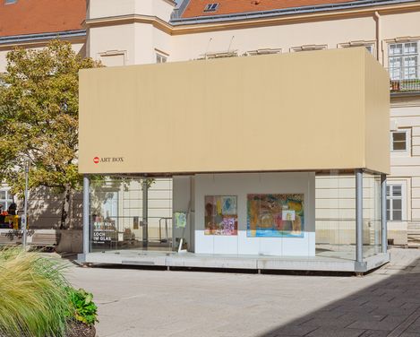 Gesamtansicht der MQ Art Box im Außenraum des MuseumsQuartier. Durch die verglaste Fassade sind mehrere farbige Arbeiten der Ausstellung sichtbar; die Box ist von historischer Architektur, Bäumen und Freiflächen umgeben.