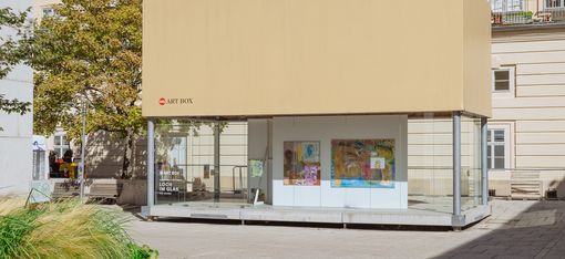 Overall view of the MQ Art Box in the outdoor area of the MuseumsQuartier. Several colorful works from the exhibition are visible through the glass façade, with historic architecture, trees, and open space surrounding the pavilion.
