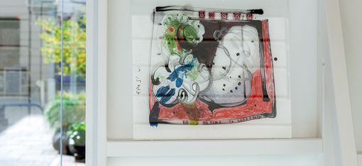 Detail view of a small-format artwork displayed within a white presentation structure. The composition combines organic, figurative forms in grey, green, blue, and red with drawn lines, small symbols, and handwritten text elements.