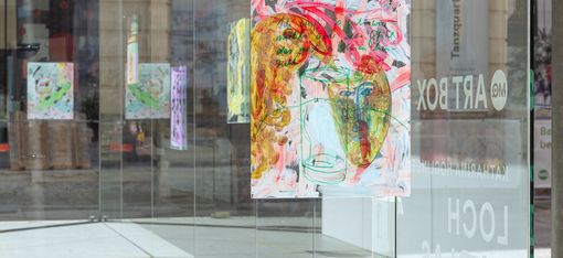 Detailansicht eines frei vor der Glasfassade hängenden, farbintensiven Bildes. Die Arbeit verbindet gestische Malerei, Zeichnung und handschriftliche Texte; weitere Werke und Spiegelungen sind im Hintergrund sichtbar.