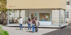 Außenansicht der MQ Art Box mit der Ausstellung „Loch im Glas“ von Katharina Höglinger. Vier Besucher:innen gehen auf die verglaste Ausstellungsfläche zu, hinter der mehrere farbenfrohe Arbeiten zu sehen sind.