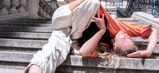 Person mit langem Haar liegt rückwärts auf einer steinernen Treppe, trägt helle Hose, orangefarbenes Oberteil und weiße Sportschuhe