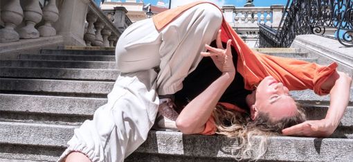 Person mit langem Haar liegt rückwärts auf einer steinernen Treppe, trägt helle Hose, orangefarbenes Oberteil und weiße Sportschuhe