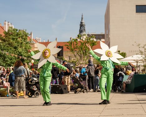Zwei Personen in grünen Overalls mit großen, weißen Blumenmasken auf dem Kopf tanzen auf einem Platz im MuseumsQuartier, umgeben von Zuschauern und Gebäuden.