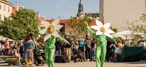 Two people wearing green overalls and large white flower headpieces dance on a plaza at MuseumsQuartier, surrounded by spectators and buildings.