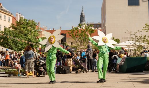 Zwei Personen in grünen Overalls mit großen, weißen Blumenmasken auf dem Kopf tanzen auf einem Platz im MuseumsQuartier, umgeben von Zuschauern und Gebäuden.