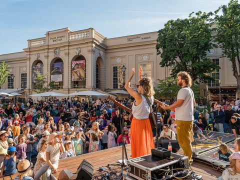 Two musicians play on a small stage in a sunny city square, crowd of families watches near a historic building.