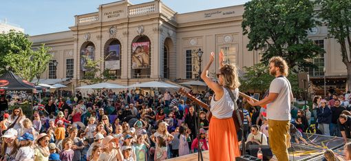 Two musicians on a stage performing to a large audience of children and adults in front of a historic building.