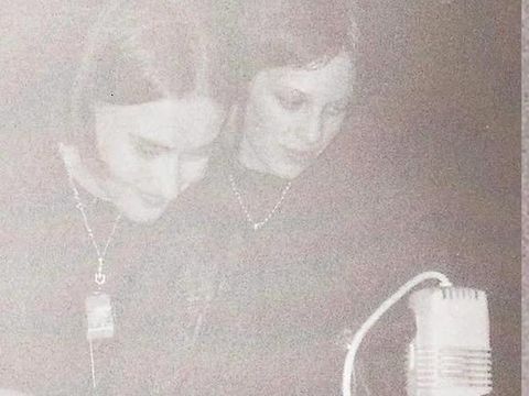 Two young women leaning over a device with a microphone in a dimly lit room