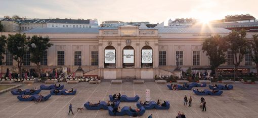 Several people are sitting and lying on curved blue seating furniture in front of a historic building with large windows and three large posters on the front of the building.