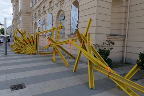 Gelbe Stahlträger mit orangen Bändern sind auf einem Gehweg vor einem hellen Gebäude zu einer abstrakten Skulptur angeordnet.