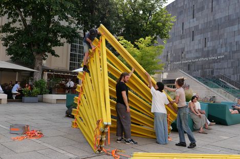 Mehrere Personen bauen im Freien eine große, gelbe, dreieckige Konstruktion aus Metallrahmen auf einem gepflasterten Platz auf