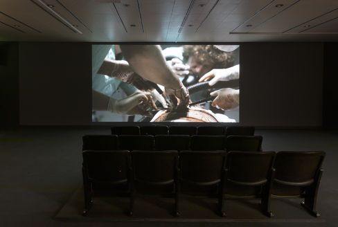 In einem dunklen Saal stehen Kinositze und auf einer Leinwand ist eine Szene aus dem Film Night Shift von Scott Clifford Evans zu sehen, in der eine Person operiert wird.