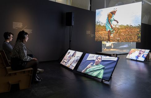 Two people wearing headphones sit in front of several tilted screens showing portraits and a large projection of a person standing in a field.