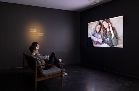 Two people sit on a sofa in a dark room watching a screen showing two other people on a sofa.