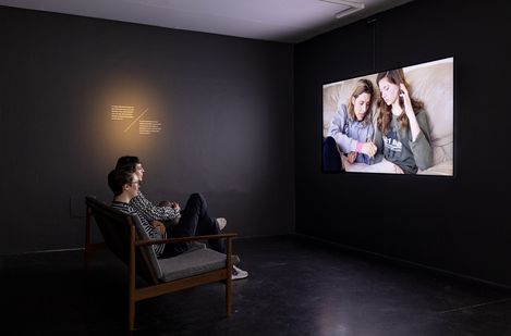 Two people sit on a sofa in a dark room watching a screen showing two other people on a sofa.