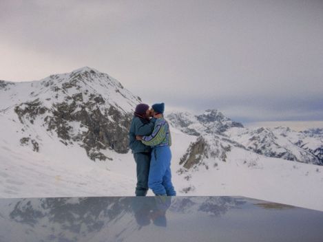 Zwei Personen in Winterkleidung umarmen sich vor schneebedeckten Bergen unter bewölktem Himmel.