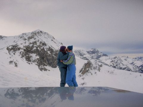 Zwei Personen in Winterkleidung umarmen sich vor schneebedeckten Bergen unter bewölktem Himmel.