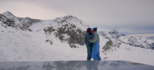 Zwei Personen in Winterkleidung umarmen sich vor schneebedeckten Bergen unter bewölktem Himmel.