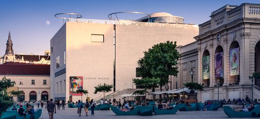 Das Leopold-Museum mit moderner und historischer Architektur, davor ein Platz mit Sitzgelegenheiten und Menschen bei Tageslicht.