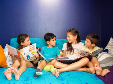 The image shows four children sitting comfortably next to each other on a large cushion. They are holding picture books and are engaged in conversation with one another. Their relaxed posture, sitting barefoot and surrounded by cushions, conveys a calm and familiar atmosphere. Overall, the image gives the impression of a protected, welcoming place where shared reading, exchange, and closeness are at the center.