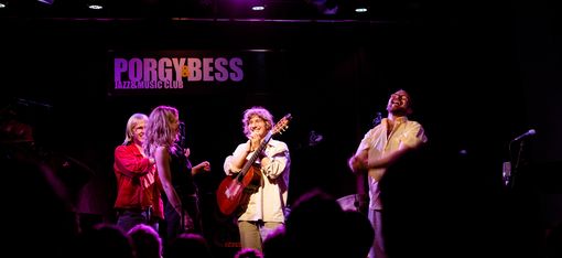 Four musicians on stage in a jazz club with one playing guitar and others singing or gesturing, audience silhouettes visible in front.