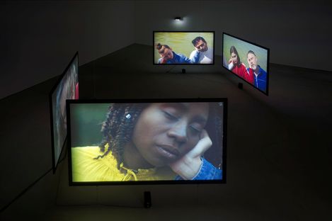Dark room with four screens showing portraits of people in various poses including a woman with closed eyes wearing a yellow top in the foreground