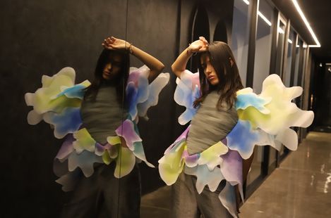 Woman with long hair and loose clothing stands in front of a mirror wearing a costume with large abstract multicolored wings.