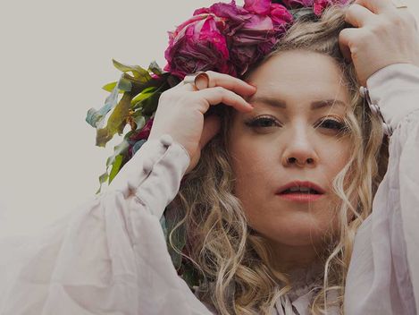 Person mit langen, lockigen Haaren, die einen Blumenkranz aus roten und rosa Blüten auf dem Kopf richtet und ein weißes, langärmeliges Kleid trägt.
