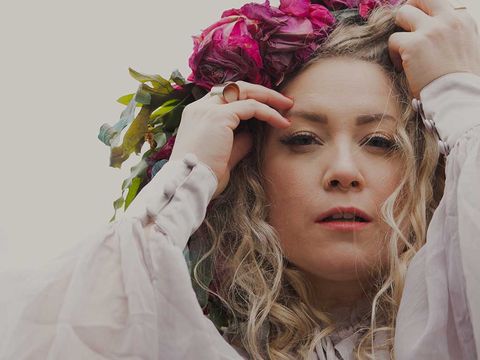 Person with long curly hair adjusting a flower crown made of red and pink blossoms while wearing a white long-sleeved dress.