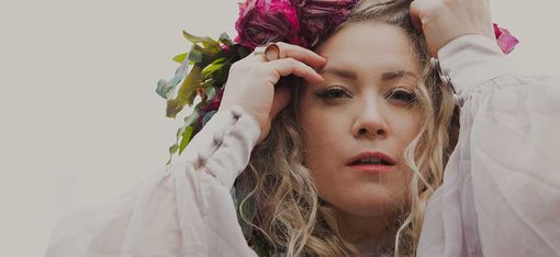 Person mit langen, lockigen Haaren, die einen Blumenkranz aus roten und rosa Blüten auf dem Kopf richtet und ein weißes, langärmeliges Kleid trägt.