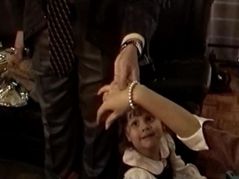 Kind mit hellem Kleid schaut nach oben und hält die Hand eines stehenden Erwachsenen, der eine Uhr und ein Armband trägt.