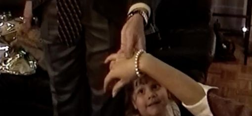 Kind mit hellem Kleid schaut nach oben und hält die Hand eines stehenden Erwachsenen, der eine Uhr und ein Armband trägt.