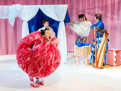 A dancer wearing a round, red costume is standing on stage. In the background, musicians are playing various instruments.