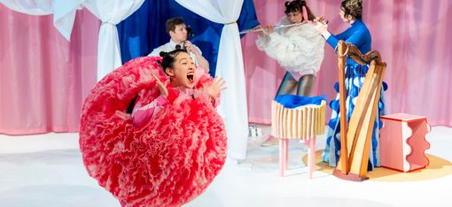 A dancer wearing a round, red costume is standing on stage. In the background, musicians are playing various instruments.