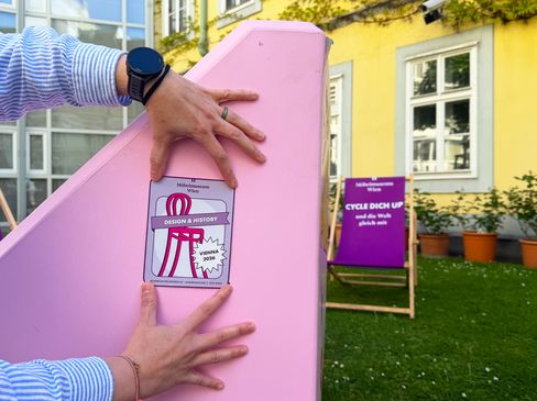 Two hands placing a “Design & History Vienna 2026” sticker on a pink Enzi; deck chairs in the background.