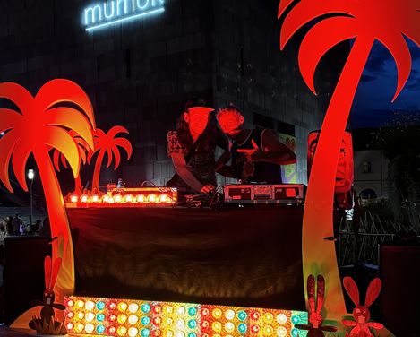Two people at a DJ booth with colorful lights and red palm tree decorations in front of a building with illuminated 'mumok' sign at night