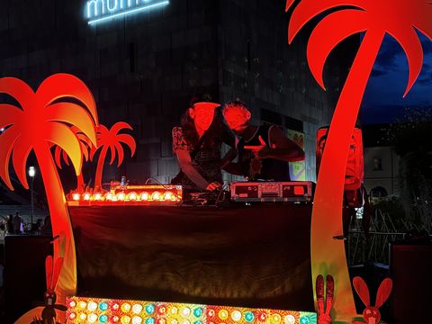 Two people at a DJ booth with colorful lights and red palm tree decorations in front of a building with illuminated 'mumok' sign at night