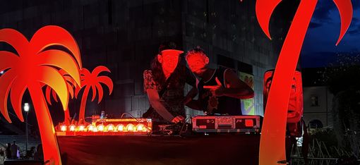 Zwei Personen an einem DJ-Pult mit bunten Lichtern und roten Palmen-Dekorationen vor einem Gebäude mit leuchtendem 'mumok'-Schriftzug bei Nacht