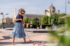 Ein kleines Mädchen mit lockigen Haaren, Sonnenbrille und blauem Kleid spielt Minigolf im Freien. Sie hält einen roten Schläger und steht vor mehreren roten Bällen auf einer hellen Bahn. Im Hintergrund sind Bäume, Spaziergänger und ein historisches Gebäude mit Kuppel zu sehen.